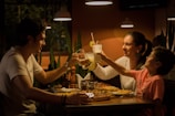 A family of three is sitting at a dining table in a warmly lit restaurant, clinking glasses for a toast. A man, a woman, and a child are smiling, enjoying a meal together. The table is set with pizza and other dishes.