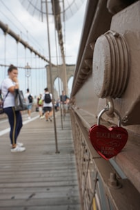 A red heart-shaped padlock engraved with names and dates hangs on a bridge railing with blurred pedestrians walking in the background.