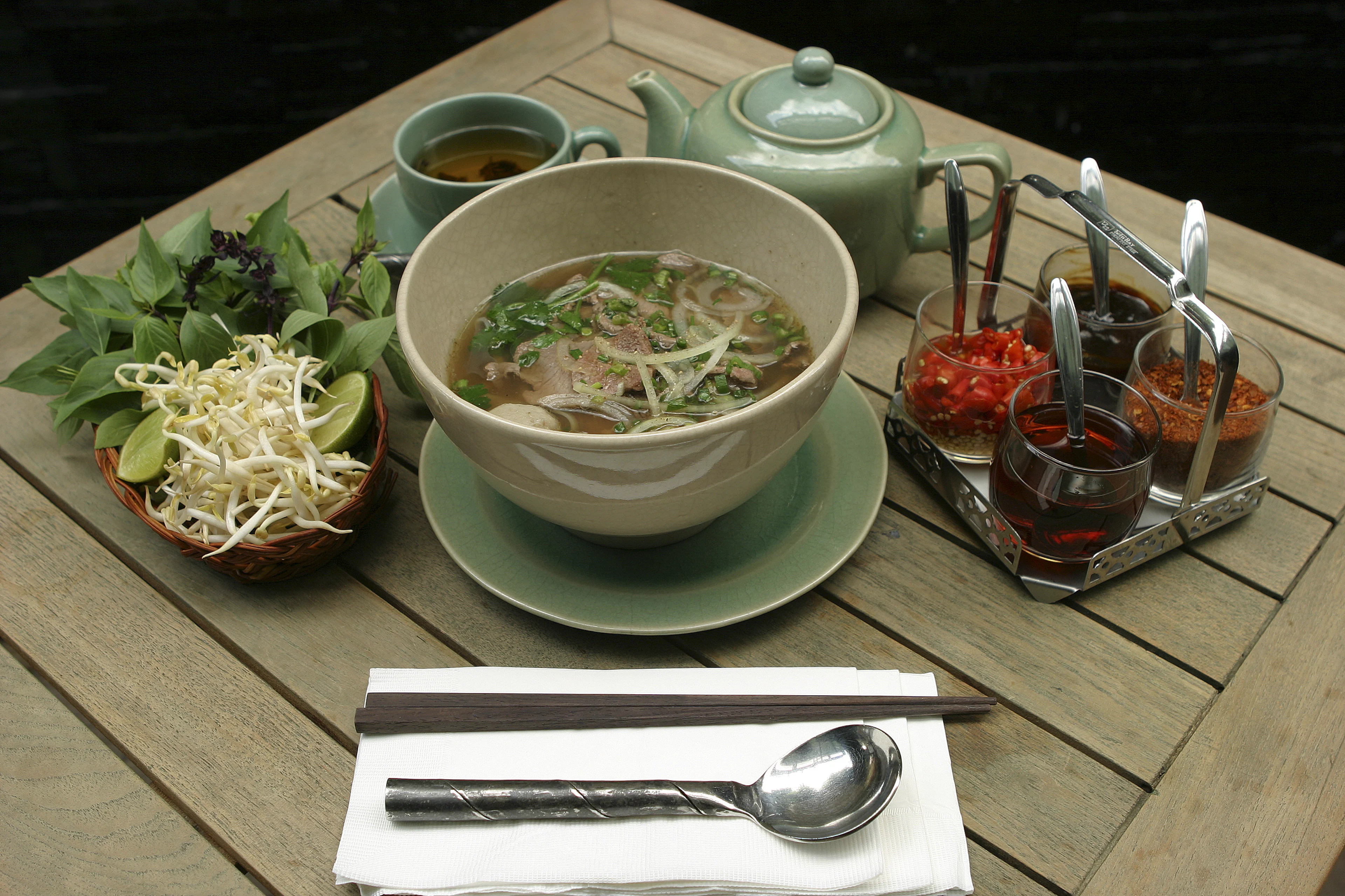 Bowl of pho on a wooden table with fresh herbs, bean sprouts and sauces