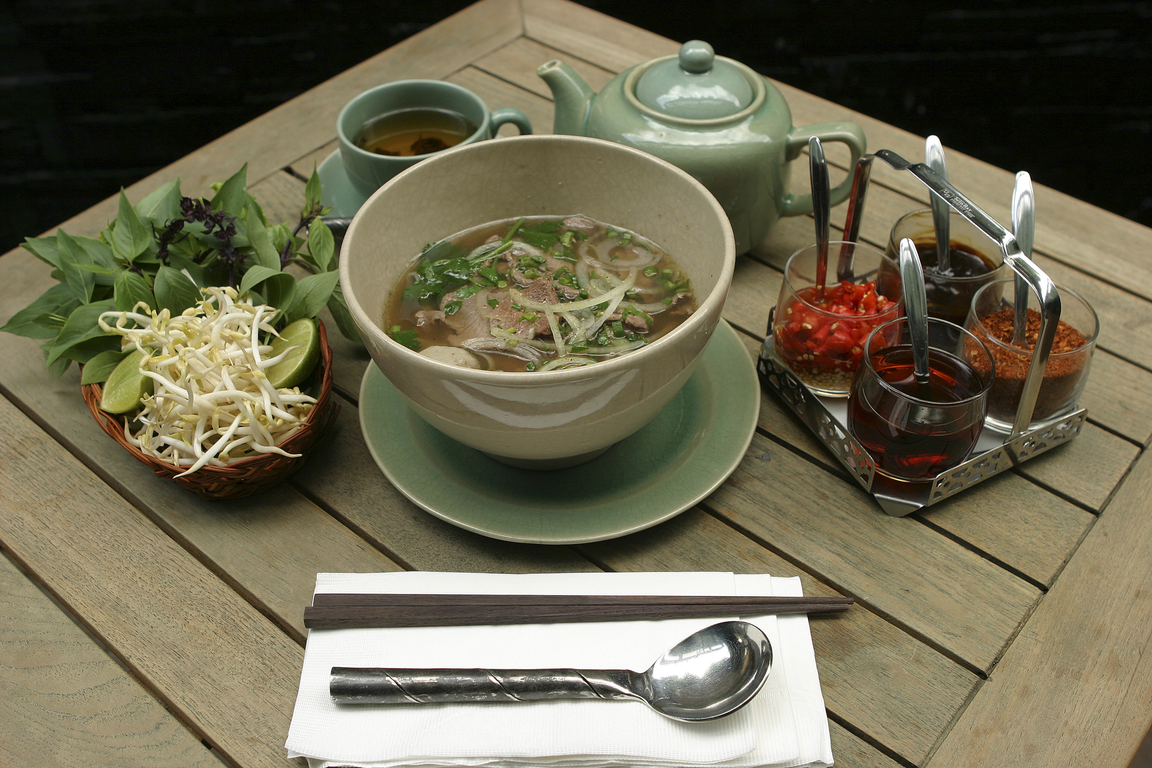 Bowl of pho on a wooden table with fresh herbs, bean sprouts and sauces