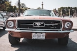 A classic Ford Mustang from 1967 is prominently featured, parked in an open area with trees and people in the background. The car has a sleek, orange exterior with a distinct grille and emblem displaying a galloping horse. The license plate is visible, and the scene is sunny with clear details of the car's front view.