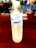 A plastic bottle of Singha drinking water with a yellow straw inside is placed on a wooden table. In the background, a person in a blue shirt is holding a smartphone.