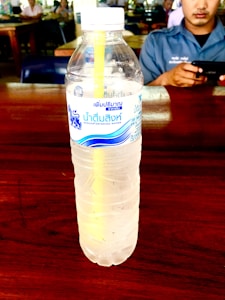 A plastic bottle of Singha drinking water with a yellow straw inside is placed on a wooden table. In the background, a person in a blue shirt is holding a smartphone.