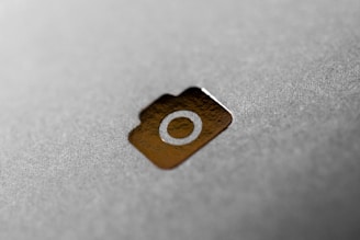 A close-up of a shiny gold camera icon embossed on a textured grey surface. The image focuses on the camera icon with a blurred background, highlighting the metallic and reflective quality of the icon.