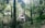 A quaint A-frame house nestled in a dense forest. Tall trees with lush green foliage surround the house, creating a serene and secluded atmosphere. The structure has a triangular roof and contrasting white and dark brown exterior panels, enhancing its rustic charm.