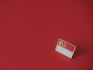 A small rectangular pin featuring the flag of Singapore is placed on a plain red surface. The flag design includes a red upper half with a white crescent moon and five stars.