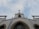 The image features a tall white church building with a prominent red cross at its peak against a partially cloudy sky. The architectural style includes pointed arch windows and decorative elements. Signs extend out from the building carrying religious words.