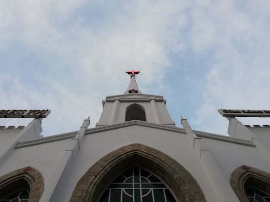 The image features a tall white church building with a prominent red cross at its peak against a partially cloudy sky. The architectural style includes pointed arch windows and decorative elements. Signs extend out from the building carrying religious words.