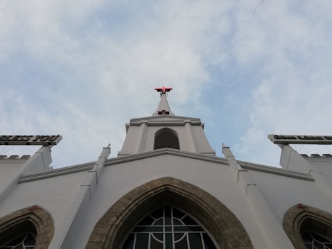 The image features a tall white church building with a prominent red cross at its peak against a partially cloudy sky. The architectural style includes pointed arch windows and decorative elements. Signs extend out from the building carrying religious words.