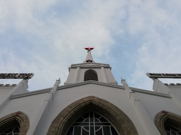 The image features a tall white church building with a prominent red cross at its peak against a partially cloudy sky. The architectural style includes pointed arch windows and decorative elements. Signs extend out from the building carrying religious words.