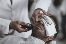 A person is lying back with a white towel covering their eyes, while someone else is applying a grooming tool to their face. The individual holding the tool wears a white garment, suggesting a professional setting like a barbershop.