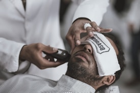 A person is lying back with a white towel covering their eyes, while someone else is applying a grooming tool to their face. The individual holding the tool wears a white garment, suggesting a professional setting like a barbershop.