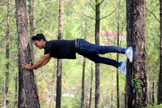 A person is horizontally suspended between two trees, wearing casual clothing and sunglasses in a wooded area.
