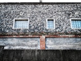 A building exterior features a unique facade of small, round stones meticulously arranged, creating a textured surface. The wall has three rectangular windows with glass blocks, providing a geometric contrast to the organic shapes of the stones. Below the stone section, there is a layer of red bricks and a worn, weathered concrete wall at the bottom.