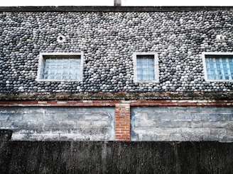 A building exterior features a unique facade of small, round stones meticulously arranged, creating a textured surface. The wall has three rectangular windows with glass blocks, providing a geometric contrast to the organic shapes of the stones. Below the stone section, there is a layer of red bricks and a worn, weathered concrete wall at the bottom.