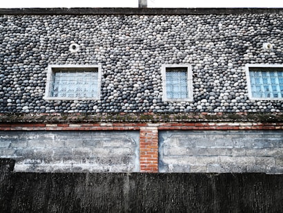 A building exterior features a unique facade of small, round stones meticulously arranged, creating a textured surface. The wall has three rectangular windows with glass blocks, providing a geometric contrast to the organic shapes of the stones. Below the stone section, there is a layer of red bricks and a worn, weathered concrete wall at the bottom.