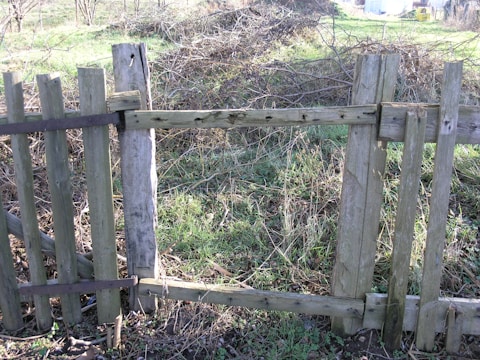 A wooden fence with missing planks stands in a field. The fence appears weathered and old, with metal brackets reinforcing some areas. Behind the fence, the ground is covered with dried grass and shrubs, indicating an untended, natural setting.