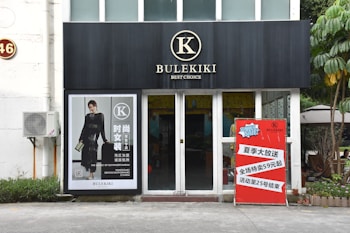 A storefront with a black sign displaying the word 'BULEKIKI' and the tagline 'Best Choice'. To the left, there is a large poster of a woman in a black dress with text in both Chinese and English. An air conditioning unit is mounted on the wall. To the right of the entrance, a red promotion sign announces a summer sale in Chinese.