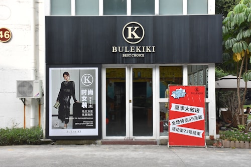 A storefront with a black sign displaying the word 'BULEKIKI' and the tagline 'Best Choice'. To the left, there is a large poster of a woman in a black dress with text in both Chinese and English. An air conditioning unit is mounted on the wall. To the right of the entrance, a red promotion sign announces a summer sale in Chinese.