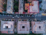An aerial view of an urban area showcasing several multi-story residential or commercial buildings. The buildings are arranged in a grid pattern with a street running horizontally through the middle of the image. Numerous cars are parked along both sides of the street. The architectural design includes flat rooftops, some with central courtyards or openings. There is greenery around the buildings, and one building has a brightly lit courtyard area.