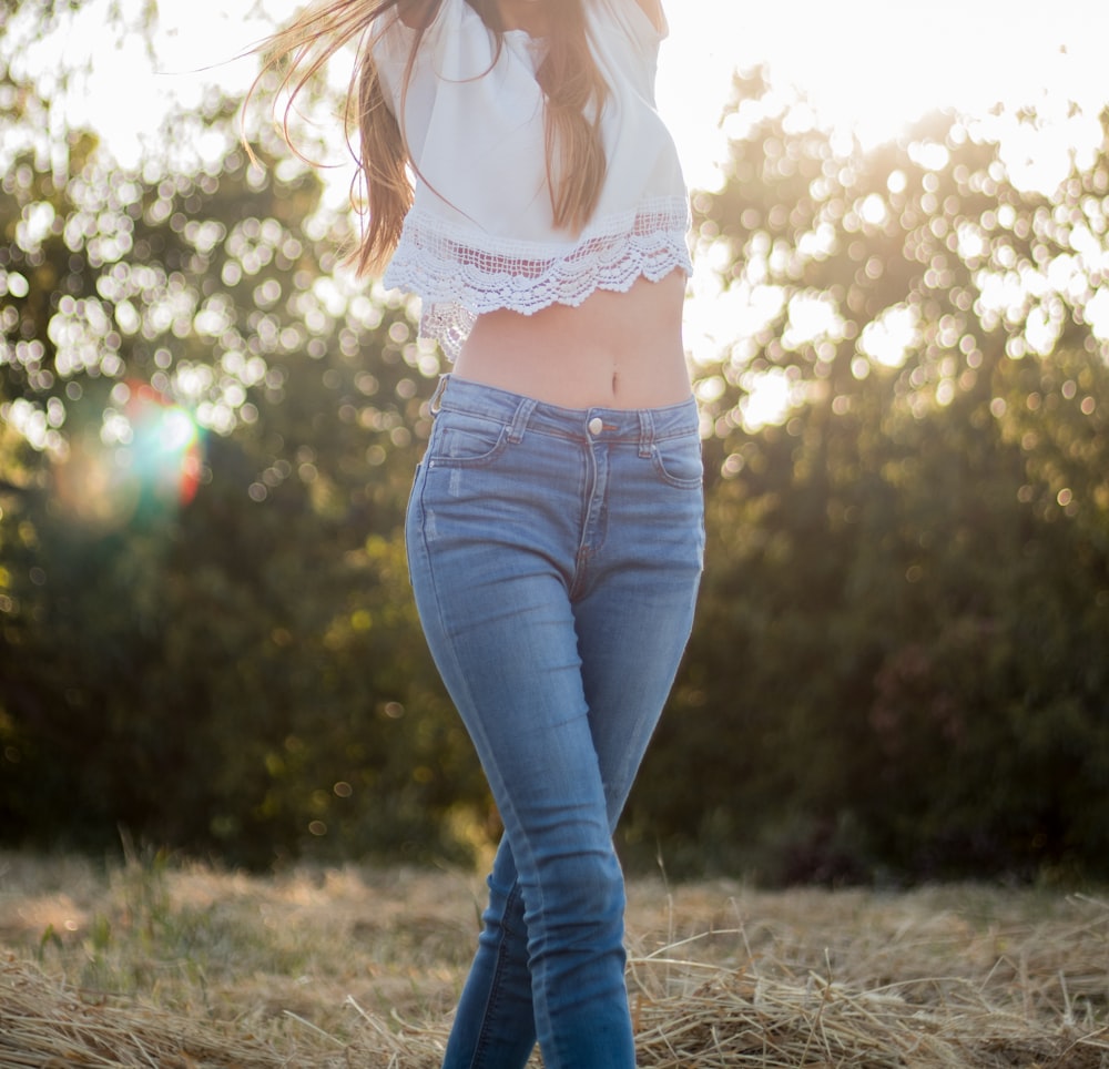 Frau trägt weißes Crop-Top und blaue Jeans, die tagsüber auf grünem Gras spazieren geht