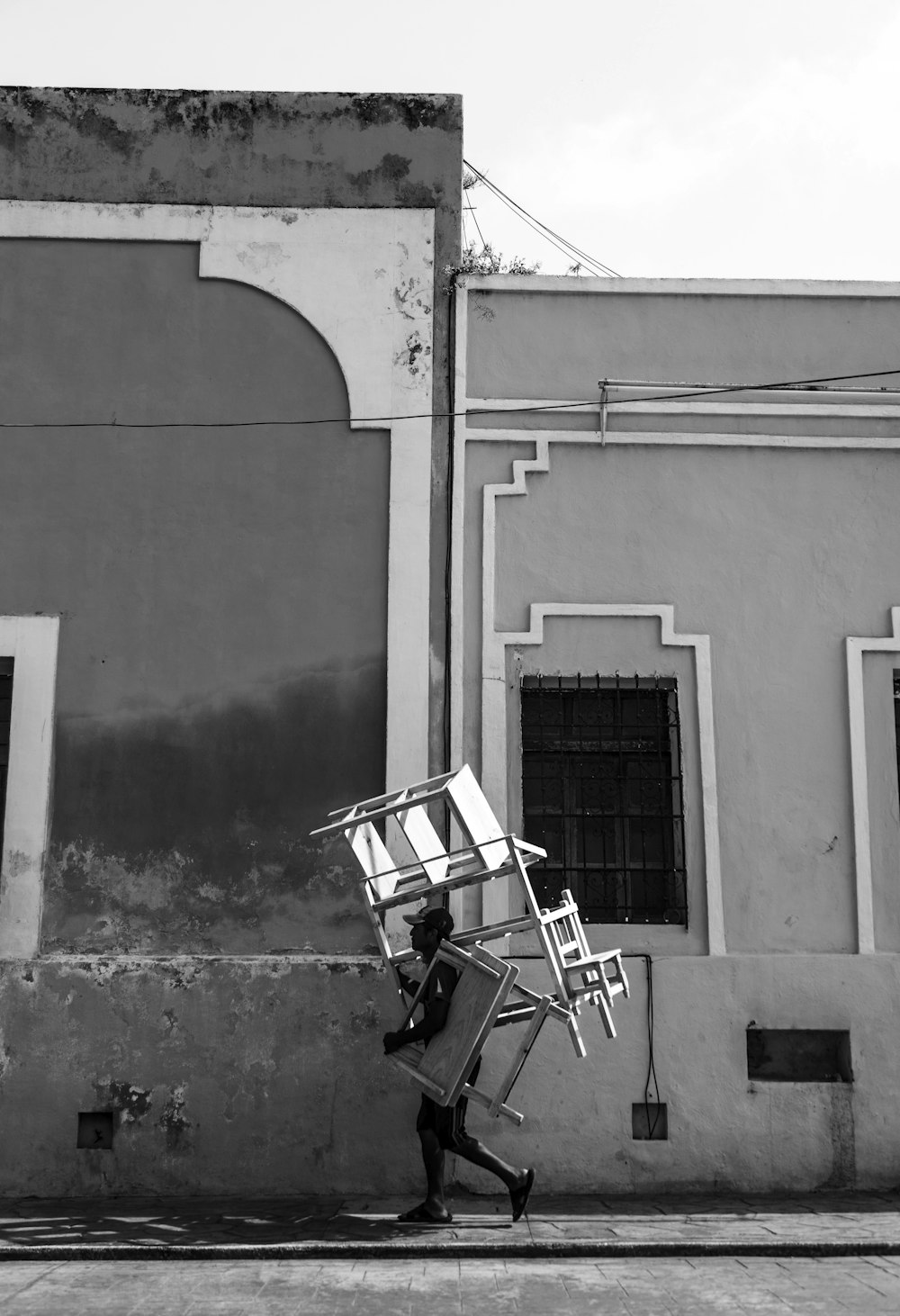 Fotografía en escala de grises de una persona que lleva muebles mientras camina cerca de una casa de hormigón durante el día