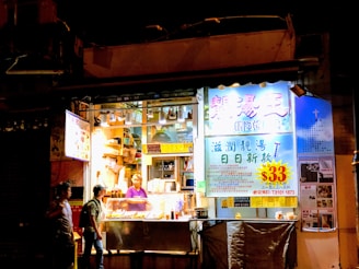 A small street food stall is brightly lit and displays a variety of food. A person inside the stall is preparing food, while another person is standing outside, seemingly placing an order. The signage is colorful, with prominent text in Chinese characters and a large price tag indicating $33.