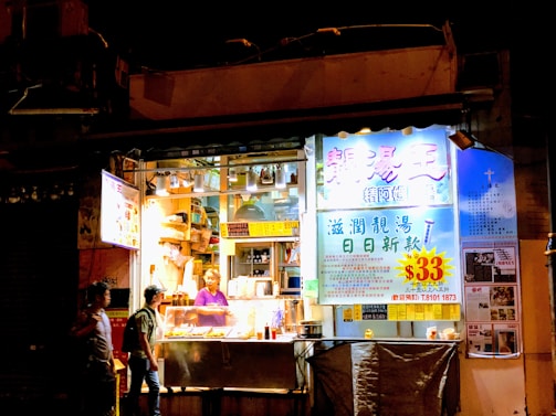 A small street food stall is brightly lit and displays a variety of food. A person inside the stall is preparing food, while another person is standing outside, seemingly placing an order. The signage is colorful, with prominent text in Chinese characters and a large price tag indicating $33.