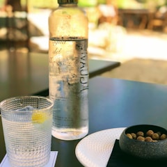 A glass bottle of water sits on a dark surface next to a clear, textured glass with a lemon wedge and ice cubes. Beside them is a small white plate and a black bowl containing olives. The background is softly blurred, suggesting an outdoor setting with warm sunlight filtering through.