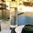 A glass bottle of water sits on a dark surface next to a clear, textured glass with a lemon wedge and ice cubes. Beside them is a small white plate and a black bowl containing olives. The background is softly blurred, suggesting an outdoor setting with warm sunlight filtering through.