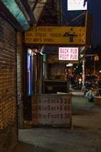 A dimly lit urban street with a massage parlor sign offering back and foot rubs. The signage is seen in yellow and red text, displaying prices and durations for services. Neon lights and a somewhat cluttered sidewalk suggest a city nightlife atmosphere.