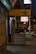 A dimly lit urban street with a massage parlor sign offering back and foot rubs. The signage is seen in yellow and red text, displaying prices and durations for services. Neon lights and a somewhat cluttered sidewalk suggest a city nightlife atmosphere.