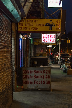 A dimly lit urban street with a massage parlor sign offering back and foot rubs. The signage is seen in yellow and red text, displaying prices and durations for services. Neon lights and a somewhat cluttered sidewalk suggest a city nightlife atmosphere.