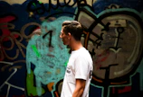 Side profile of a young man in a minimalist white tee, walking past a graffiti wall.