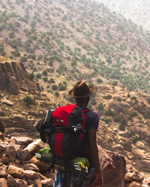 A person wearing a hat and carrying a large red backpack stands on rocky terrain, gazing towards a mountainous landscape dotted with sparse vegetation. The scene suggests a rugged outdoor adventure.