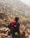 A person wearing a hat and carrying a large red backpack stands on rocky terrain, gazing towards a mountainous landscape dotted with sparse vegetation. The scene suggests a rugged outdoor adventure.