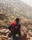 A person wearing a hat and carrying a large red backpack stands on rocky terrain, gazing towards a mountainous landscape dotted with sparse vegetation. The scene suggests a rugged outdoor adventure.