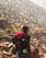 A person wearing a hat and carrying a large red backpack stands on rocky terrain, gazing towards a mountainous landscape dotted with sparse vegetation. The scene suggests a rugged outdoor adventure.