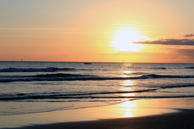 A tranquil beach scene during sunset with gentle waves rolling onto the shore. The sun is low on the horizon, casting a golden glow across the water and sky, with some scattered clouds visible.