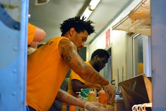 Two individuals are seen working inside a food truck, wearing orange shirts. One person is preparing food with gloved hands, placing items into a basket, while various sauce bottles are visible on the counter. The inside of the truck has shelves and bright lighting.