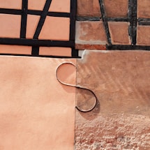 A segment of a building wall that displays a mix of textures and colors. The wall features two distinct plaster sections, one with a smooth peach tone and the other with a rougher, darker beige surface. Black wooden beams form a geometric pattern on the upper section. A curved metal rod resembling an 'S' shape is attached, adding an artistic element.