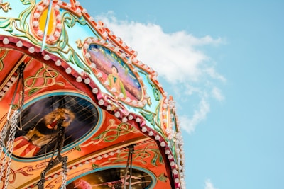 A brightly colored carousel with intricate patterns and paintings on its canopy. The carousel is captured with a backdrop of a clear blue sky with a few clouds. Vibrant decorative elements include flowers, ribbons, and figures.