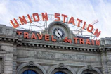 The facade of a historic train station features large, prominent signage. An ornate clock is positioned centrally above the entrance, surrounded by classical architectural detailing. The words 'Union Station' and 'Travel by Train' are displayed in illuminated letters, with a crane visible in the background.