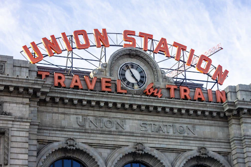 The facade of a historic train station features large, prominent signage. An ornate clock is positioned centrally above the entrance, surrounded by classical architectural detailing. The words 'Union Station' and 'Travel by Train' are displayed in illuminated letters, with a crane visible in the background.