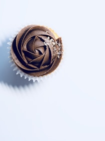 A chocolate cupcake with a swirl pattern is topped with edible glitter. The cupcake sits in a white paper liner against a plain, light background. Shadows are cast to the left, adding depth.