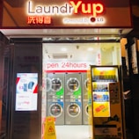 A brightly lit laundromat is open for use, featuring a row of washing machines. The storefront has signs in both English and Chinese, with a vending machine on the right and a digital advertisement on the left. The flooring is white with a yellow caution sign indicating a wet floor.