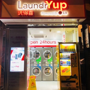 A brightly lit laundromat is open for use, featuring a row of washing machines. The storefront has signs in both English and Chinese, with a vending machine on the right and a digital advertisement on the left. The flooring is white with a yellow caution sign indicating a wet floor.