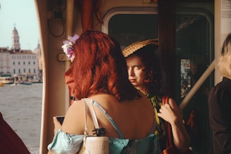 Two women with red hair are on a boat, with one facing away and the other looking directly at the camera. The one facing away has a flower in her hair, while the other wears a straw hat. The background reveals a view of a waterfront with historical buildings.