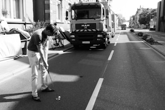 A person is playing golf on a street, with a large truck parked nearby and buildings lining the road. A bicycle and some debris can be seen on the sidewalk.