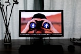 A computer monitor displays an image of a person looking through binoculars with Facebook logos on the lenses. The setup includes a desktop and a glass vase with long, dark plant stems on a wooden table, with curtains in the background.